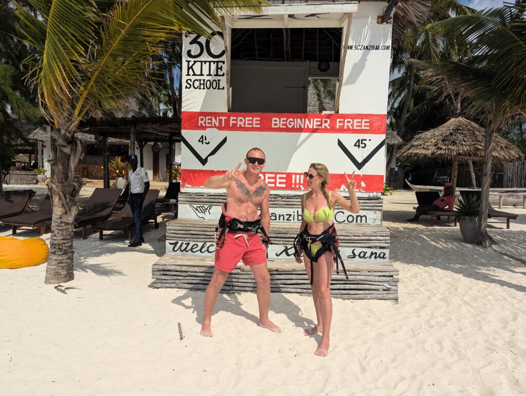 An instructor guiding a beginner kitesurfer at 3C Zanzibar kitesurf school on Paje Beach, with kites flying overhead.